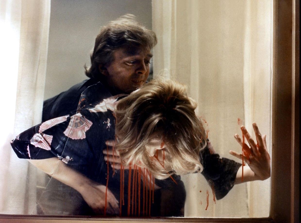 Marcus tenant le corps de Helga Ulmann en sang, les mains plaquées sur la vitre de sa fenêtre
